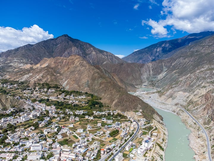 An aerial view of a mountain town nestled between two mountains, with a winding river running through the valley. The town is surrounded by lush greenery and houses, and the sky is a clear blue.