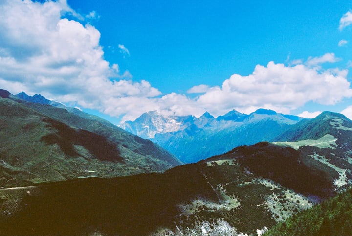 A mountainous landscape with lush greenery, a winding road, and a clear blue sky with fluffy white clouds.