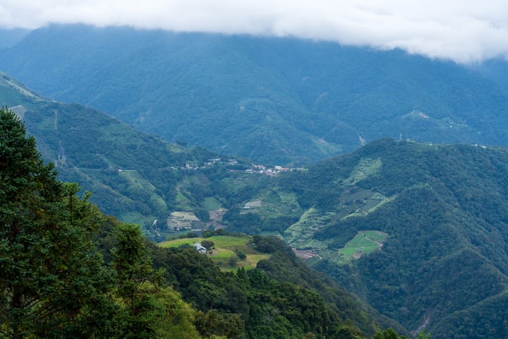 A serene mountain landscape with lush greenery, a small village nestled among trees, and a cloudy sky.
