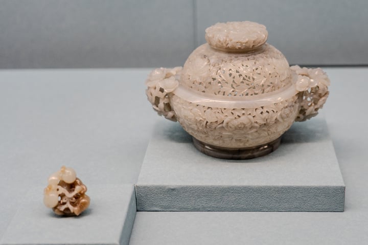 A beige pottery vase with a round base and two handles is displayed on a gray platform, with a small, brown, and white gemstone on the right side.