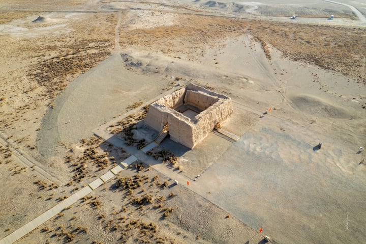 An aerial view of a desert area reveals a large, rectangular stone structure with a square base, surrounded by sparse vegetation. The structure is centrally located, with a dirt path leading to it. The sky is clear, and the ground is dry, suggesting a hot climate.