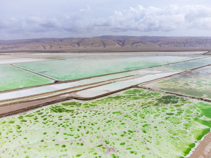 An aerial view of a vast, green lake surrounded by brown mountains and a cloudy sky, with a few scattered clouds.