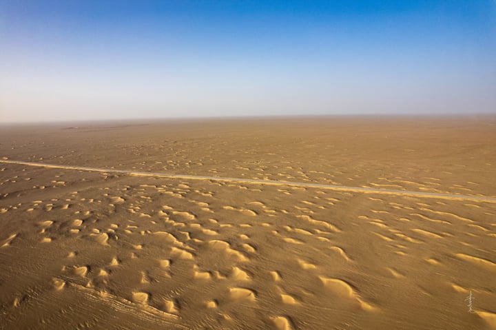 An aerial view of a vast, sandy desert with a winding dirt road, creating a pattern of small circular shapes, and a clear blue sky above.
