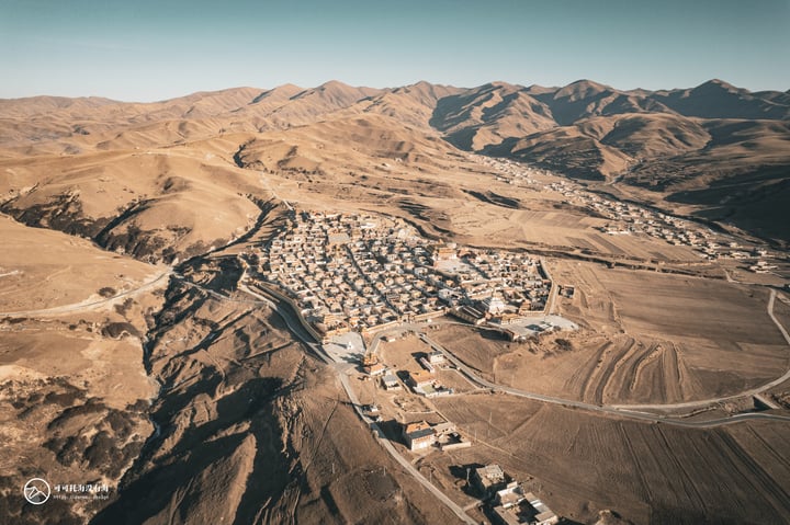 An aerial view of a small town nestled in a valley, surrounded by mountains and surrounded by a vast expanse of brown dirt. The town is densely packed with white buildings and roads, and the sky is a clear blue.