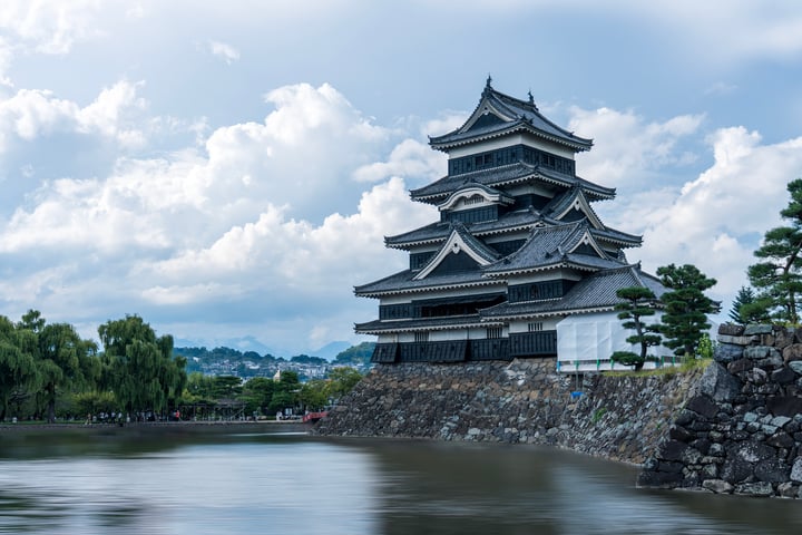 A majestic, multi-level Japanese castle stands on a stone wall, surrounded by a serene body of water. The sky is a clear blue with white clouds, and the castle is set against a backdrop of lush green trees.