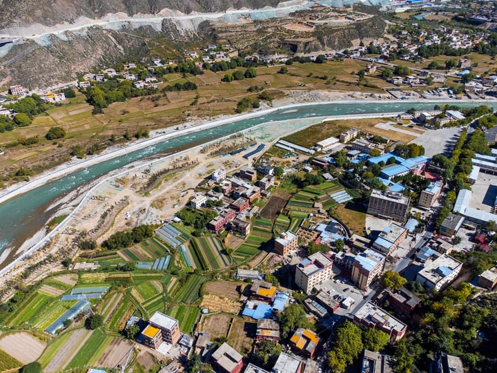 An aerial view of a village nestled between a river and mountains, with buildings of varying colors and structures, surrounded by green fields and trees.