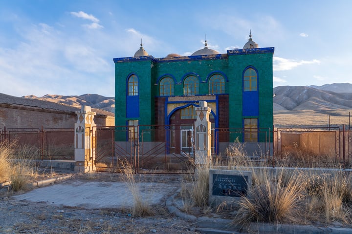A blue and green church with two domes stands in a desert-like landscape, surrounded by a fence and a monument. Mountains are visible in the distance.