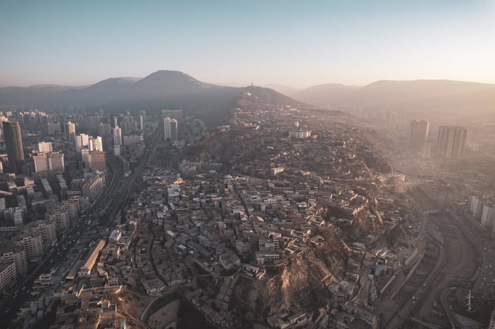 An aerial view of a mountainous city with a hillside, densely populated with buildings of varying heights, surrounded by greenery. The city is situated on a hill, with roads and highways running through it. The sun casts a warm glow on the city, highlighting its architectural details.
