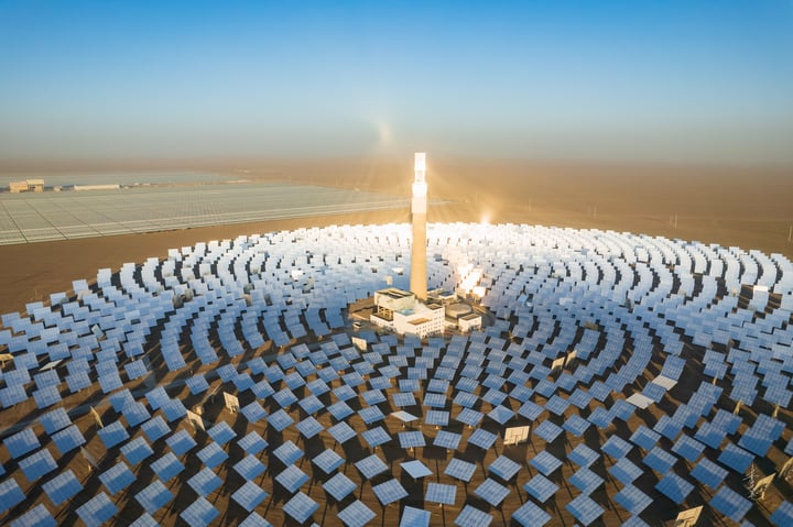 An aerial view of a large solar farm in a desert-like environment, with solar panels arranged in a circular pattern and a large white sun tower in the center. The farm is surrounded by a vast expanse of sandy terrain and a few wind turbines in the distance.