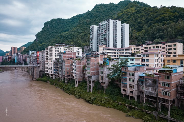 A cityscape with tall, white and gray buildings, some with balconies, is nestled on a riverbank, with a bridge connecting the two sides. The river is brown and flowing, and the sky is cloudy.