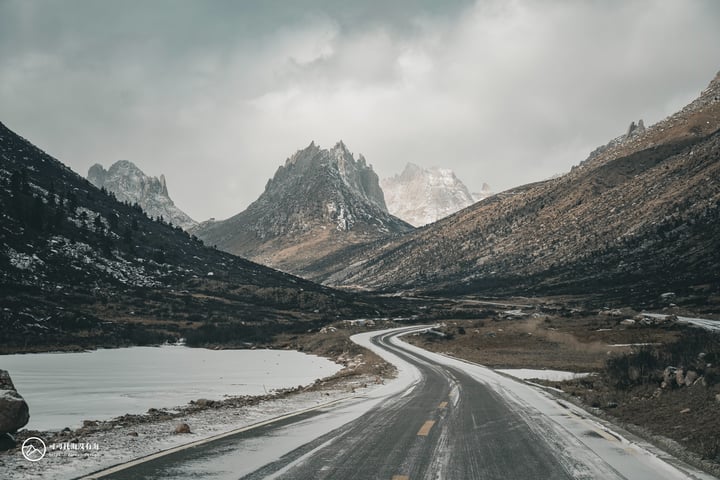 A winding, wet road winds through a mountainous landscape, with snow-capped mountains in the background and a body of water nearby. The sky is cloudy, casting a soft light over the scene.