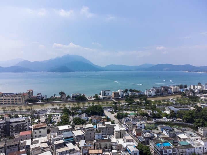 A panoramic view of a coastal city with a large body of water, boats, and mountains in the background.
