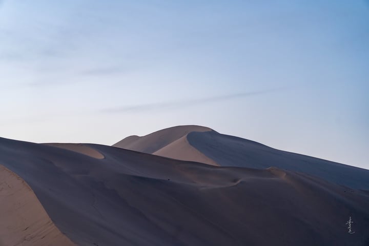 A desert landscape with sand dunes, a clear blue sky, and a solitary figure on a hill.