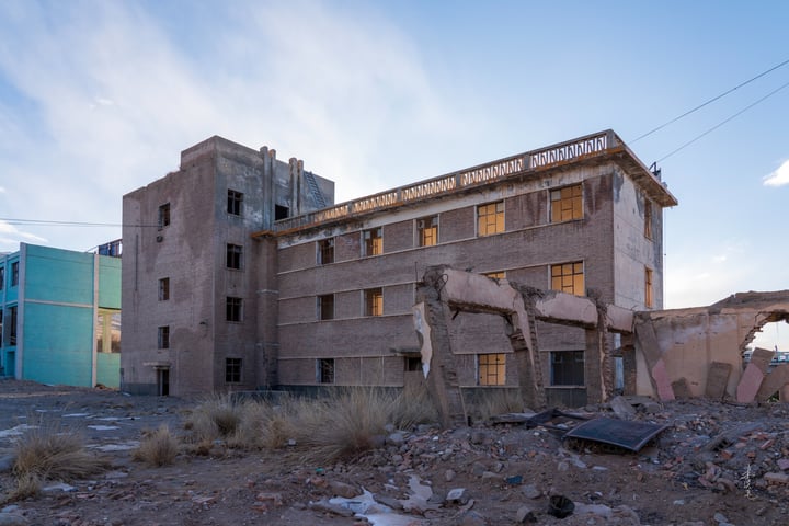 An old, dilapidated building with a large hole in the middle, surrounded by debris and power lines, set against a clear blue sky.