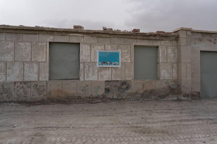 An old, dilapidated building with a blue sign advertising "Pens" on its side, surrounded by a dirt road and a cloudy sky.