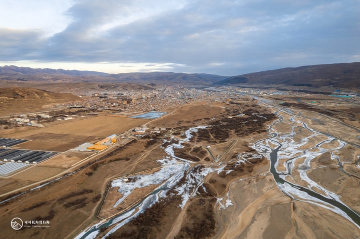 An aerial view of a mountainous landscape with a winding river, surrounded by buildings and a cloudy sky.