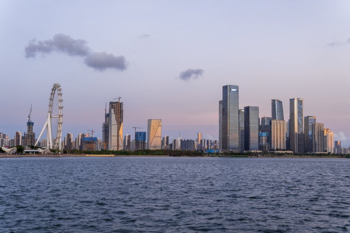 A serene cityscape is captured with a large body of water in the foreground, featuring a white Ferris wheel and a few other buildings, all set against a pale blue sky.