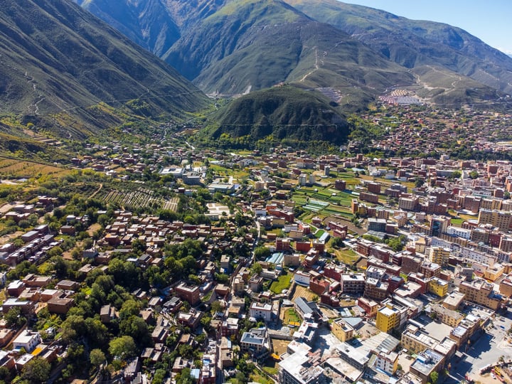 An aerial view of a mountainous town with red-roofed buildings, green spaces, and a clear blue sky.