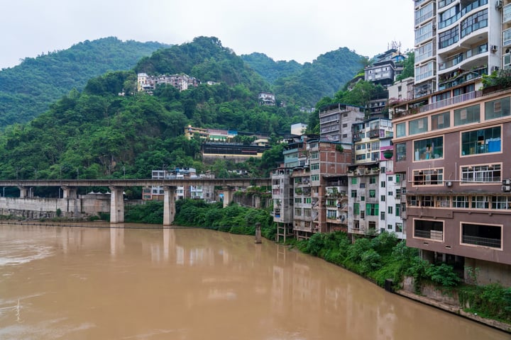 A serene cityscape with a brown river, a bridge, and a hillside with green trees and buildings.