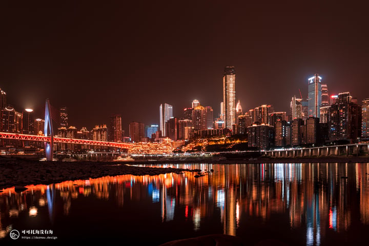 A nighttime cityscape is reflected in calm water, with tall buildings illuminated by lights and a bridge crossing the river.