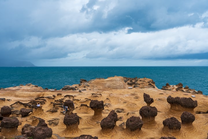 A rocky coastline with sand dunes and small rock formations, overlooking a deep blue ocean and a cloudy sky.