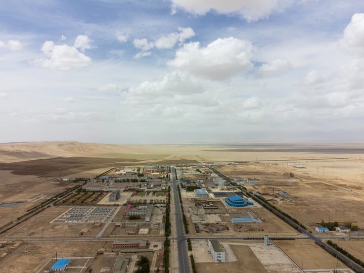 An aerial view of a large, complex, and densely populated area with a mix of residential and commercial structures, surrounded by a vast expanse of barren, sandy terrain. The sky is a clear blue with scattered clouds.