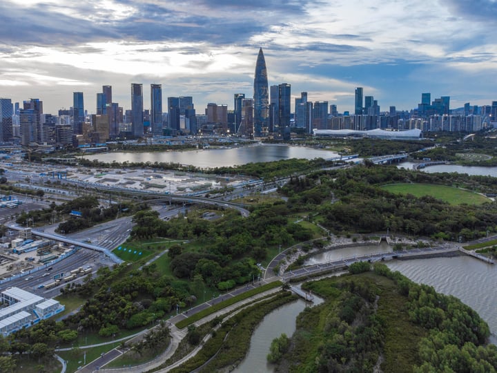 An aerial view of a city with a skyline of tall buildings, a river, and green spaces, with a cloudy sky above.