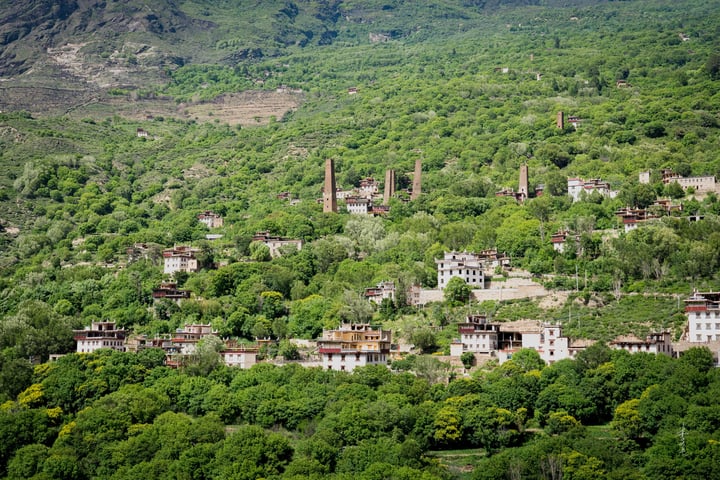 A serene mountainous landscape features a quaint village nestled amidst lush greenery, with white and red buildings scattered across the hillside. The village is surrounded by trees, and the sky is a clear blue.