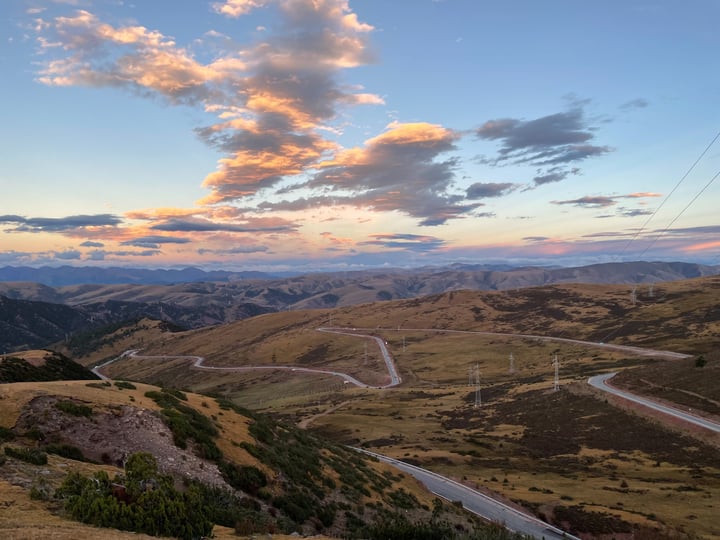 A winding mountain road winds through a valley, surrounded by rolling hills and a cloudy sky. The road is surrounded by grassy areas and power lines, creating a serene and picturesque scene.