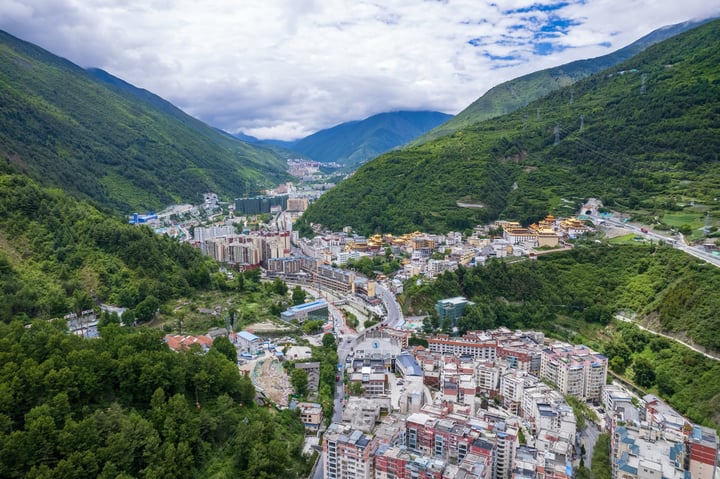 An aerial view of a mountain town with a winding road, surrounded by lush green mountains and a blue sky with clouds.