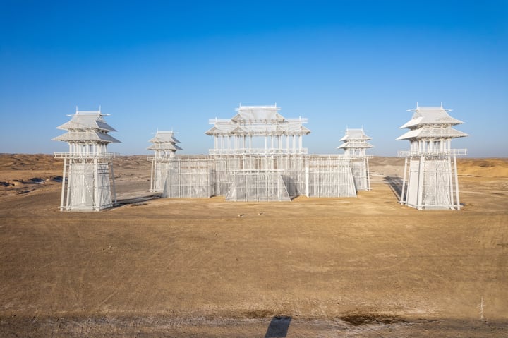 An aerial view of a white, pagoda-like structure with multiple levels and a central roof, surrounded by a barren desert landscape.
