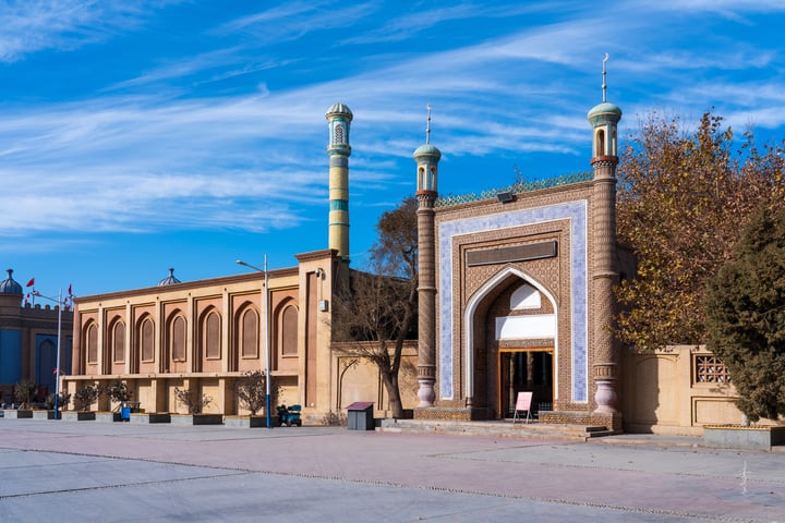 A serene scene features a large, ornate archway with blue and white tiles, flanked by two green minarets and a blue dome. The archway is set against a backdrop of a clear blue sky with scattered clouds. The archway is surrounded by lush green trees and a fence, creating a tranquil atmosphere.