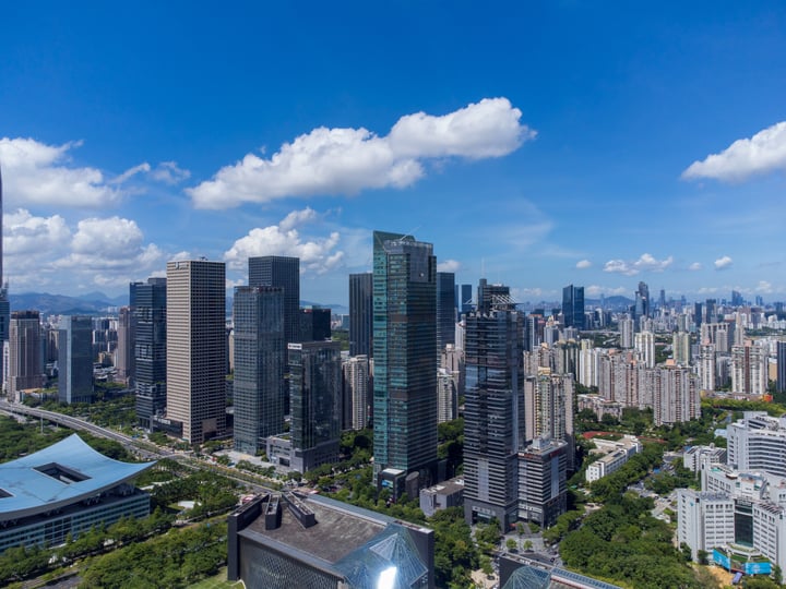 The image showcases a bustling cityscape with tall buildings, some of which have a modern design, and others with a more traditional look. The buildings are scattered across the city, with some closer to the viewer and others further away. The sky above is a clear blue with a few clouds, and the city appears to be in a high-rise building area. The image captures the essence of urban life, with the city's architecture and the sky's color contrasting.