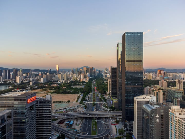 A cityscape at sunset, with a mix of modern and older buildings, a large highway, and a river, surrounded by mountains.