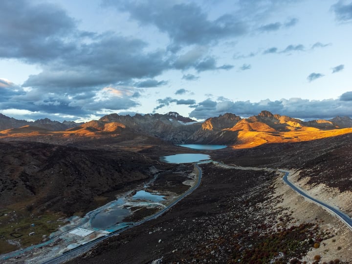 A scenic mountainous landscape with a winding road, a mountain range, and a lake in the foreground.