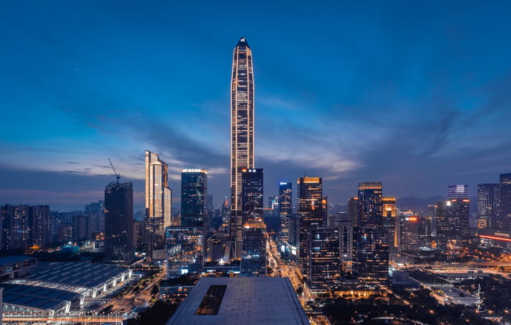 A nighttime cityscape features a tall, curved skyscraper with a pointed top, illuminated by warm lights, surrounded by other tall buildings and a busy road. The sky is dark blue with scattered clouds, and the city lights create a vibrant contrast.