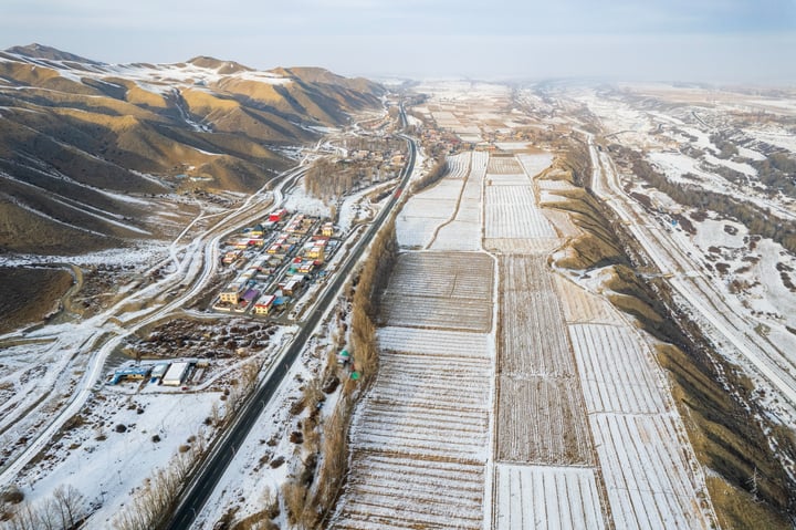 An aerial view of a snowy landscape with a winding road, surrounded by mountains and houses, with a clear blue sky above.