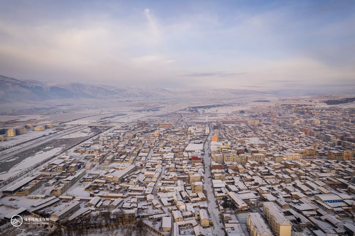 An aerial view of a snowy city with a mix of buildings and roads, surrounded by mountains and a clear blue sky.
