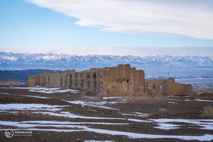 An old stone building with a doorway and windows stands in a barren landscape, surrounded by snow-covered mountains and a clear blue sky.