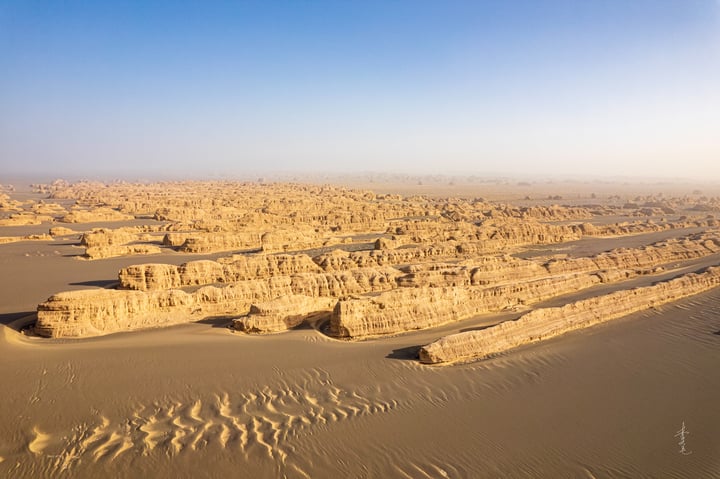 Aerial view of a desert landscape with sandy formations, a clear blue sky, and a few distant structures.