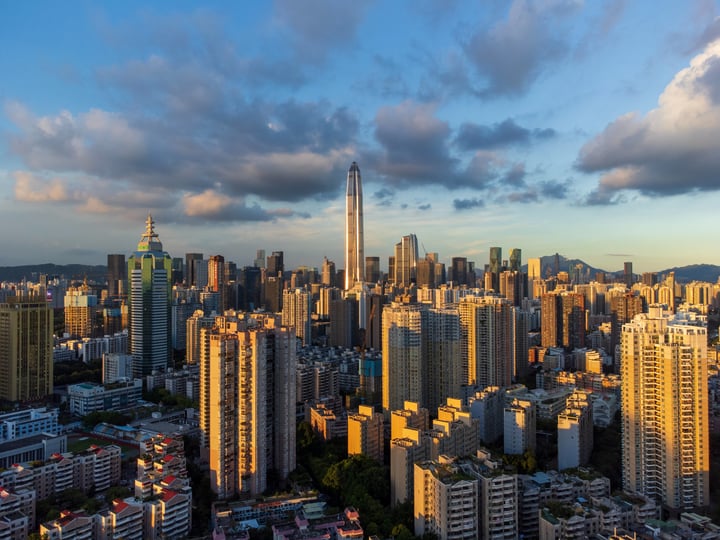 A panoramic cityscape at sunset, with a mix of modern and older buildings, a tall skyscraper, and a mountain in the distance.