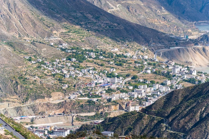 A mountainous valley with a winding road, white buildings, and green vegetation, with a clear blue sky above.