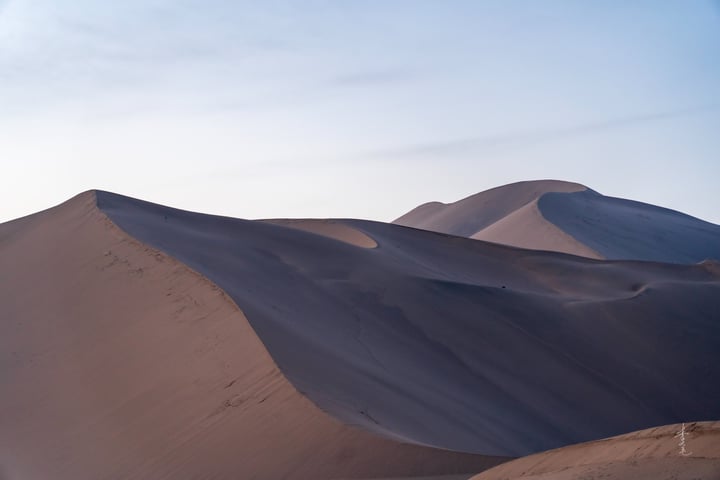 A desert landscape with sand dunes, a clear blue sky, and a lone white pole in the foreground.