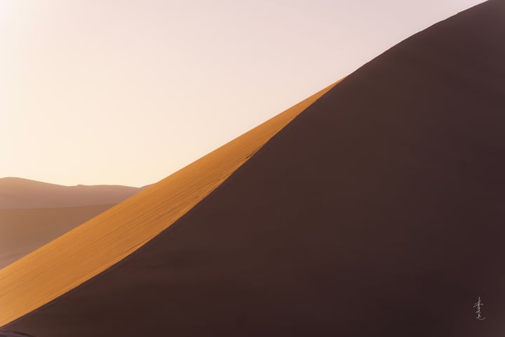 A solitary figure stands on a sandy hill in a vast desert landscape, with a clear blue sky above.