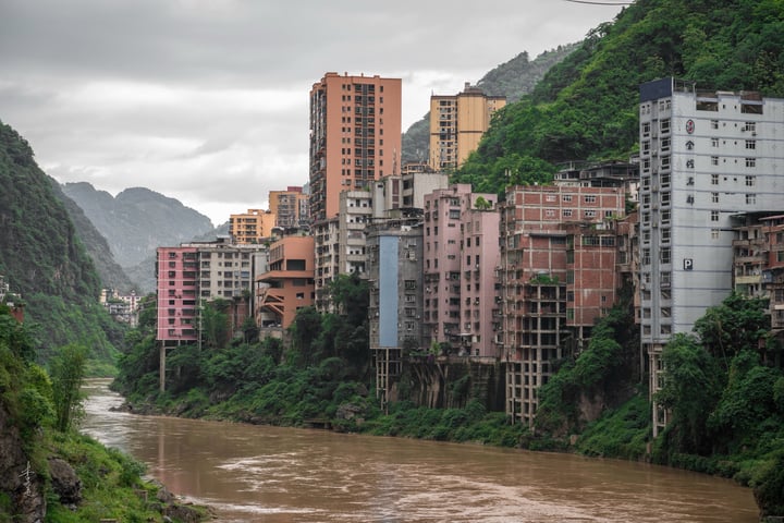 A cityscape with tall buildings on a riverbank, surrounded by mountains and greenery, with a cloudy sky overhead.