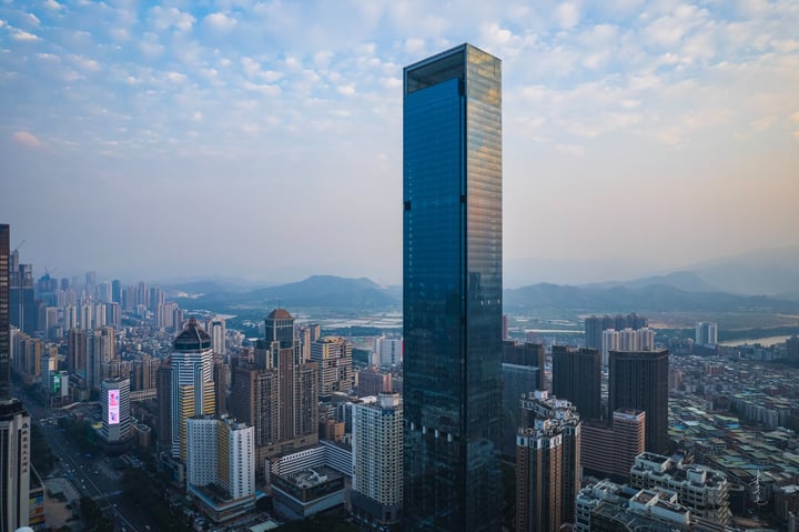 A high vantage point captures a bustling cityscape with a prominent skyscraper, surrounded by other buildings and a river. The sky is a mix of blue and pink hues, and mountains are visible in the distance.