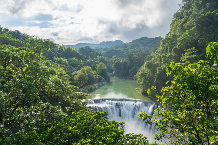 A serene landscape features a waterfall cascading down a mountain, surrounded by lush greenery and a clear blue sky with white clouds.