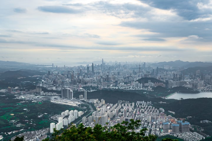A panoramic cityscape with a mix of modern and traditional buildings, surrounded by mountains and a body of water, is captured in a high-angle shot.