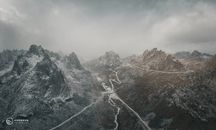 A snowy mountain range with a winding road through it, surrounded by a cloudy sky.
