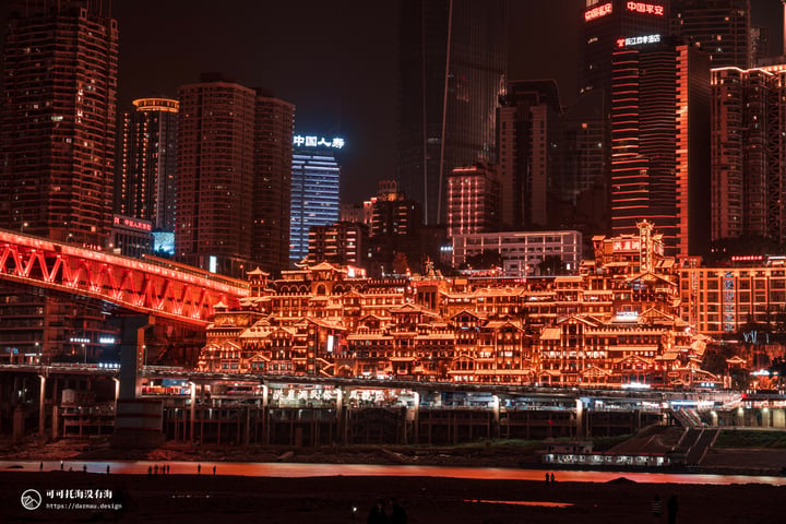 A nighttime cityscape is reflected in calm water, with tall buildings illuminated by lights and a bridge crossing the river.
